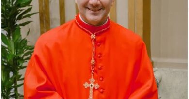 Cardinal in bright red ceremonial robes and a red zucchetto, standing indoors with hands folded and a cross necklace visible.