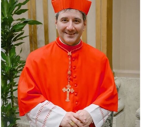 Cardinal in bright red ceremonial robes and a red zucchetto, standing indoors with hands folded and a cross necklace visible.