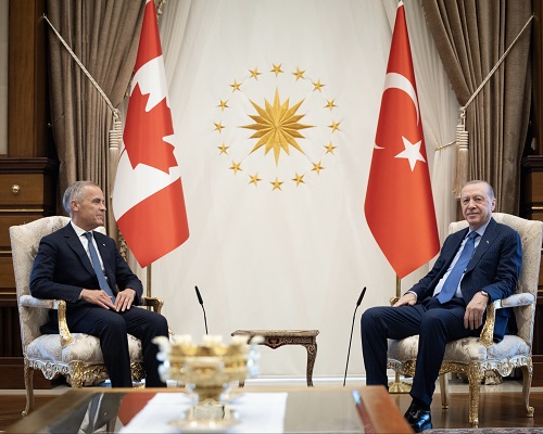Two world leaders sit in ornate chairs for a formal diplomatic meeting, with Canadian and Turkish flags behind them and a sun emblem in the center.