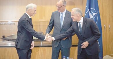 Three men in suits shake hands in a formal meeting room, with a NATO flag visible in the background and a model display case nearby.