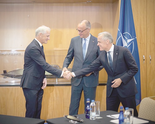 Three men in suits shake hands in a formal meeting room, with a NATO flag visible in the background and a model display case nearby.