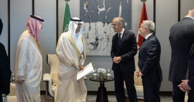 Two men in traditional Middle Eastern attire and two suited officials exchange documents in a formal meeting room with flags and a modern painting behind them.