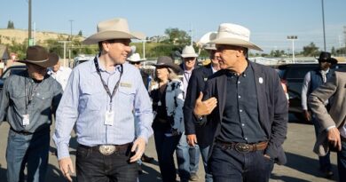 Group of cowboys walking outdoors, two men in hats chat while others follow, in a parking lot on a sunny day with lanyards.
