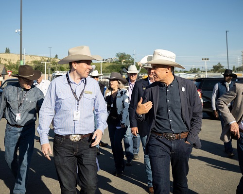 Group of cowboys walking outdoors, two men in hats chat while others follow, in a parking lot on a sunny day with lanyards.