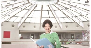 Woman in a green sweater holds a blue folder and smiles in a bright indoor atrium with a glass dome roof.