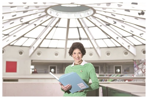 Woman in a green sweater holds a blue folder and smiles in a bright indoor atrium with a glass dome roof.