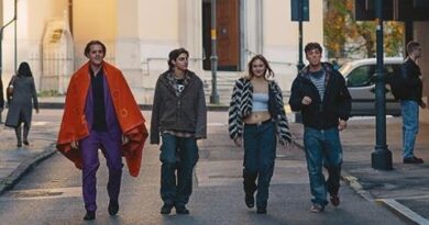 Four young adults walk down a city street; the leftmost man wears a bright orange cape and purple pants, others in casual jackets as they stroll.