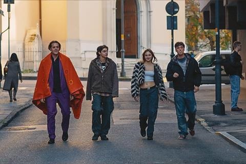 Four young adults walk down a city street; the leftmost man wears a bright orange cape and purple pants, others in casual jackets as they stroll.