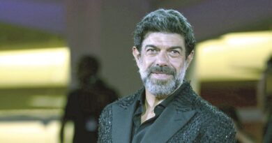 Bearded man in a dark, sparkly jacket smiles at the camera at an indoor event.