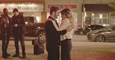 A couple embraces on a neon-lit city sidewalk at night, with two men chatting in the background near storefronts.