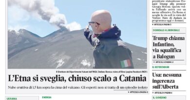 Man in a dark jacket standing on snow on Mount Etna with smoke rising from the crater behind him.