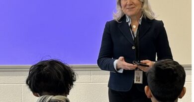 Teacher in a navy blazer speaks to students in a classroom, standing at the front by a purple board.