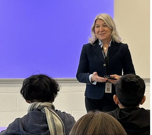 Teacher in a navy blazer speaks to students in a classroom, standing at the front by a purple board.