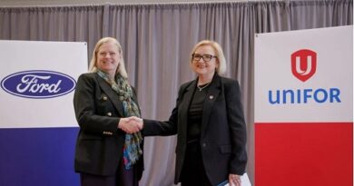 Two women in dark business suits shake hands on a stage, with Ford and UNIFOR banners in the background.