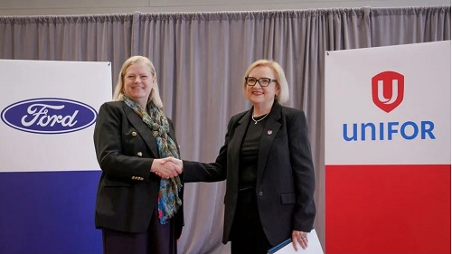 Two women in dark business suits shake hands on a stage, with Ford and UNIFOR banners in the background.
