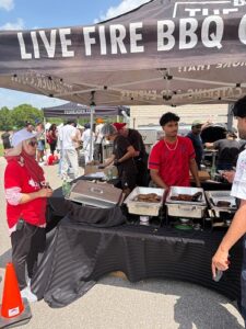 Outdoor Live Fire BBQ stall under a black tent with a large 'LIVE FIRE BBQ' banner; staff grill and trays of meat with attendees nearby.
