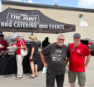 Two men smile under a BBQ catering tent labeled 'The Joint,' with several people serving food in the background.