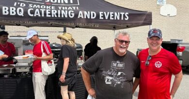 Two men smile under a BBQ catering tent labeled 'The Joint,' with several people serving food in the background.