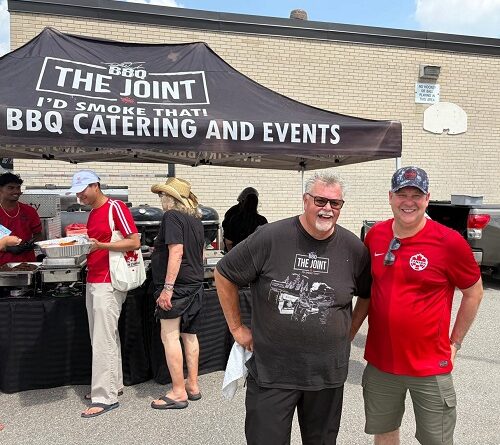Two men smile under a BBQ catering tent labeled 'The Joint,' with several people serving food in the background.