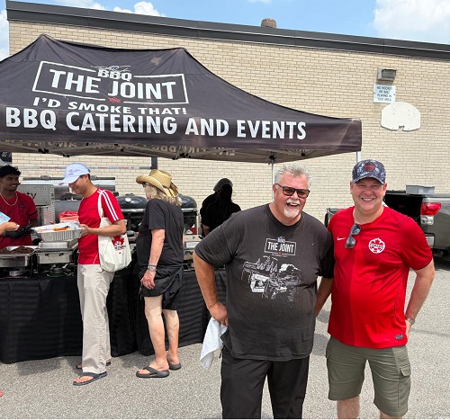 Two men smile under a BBQ catering tent labeled 'The Joint,' with several people serving food in the background.