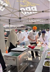 Outdoor food stall under a canopy where a diverse group of people, some wearing turbans, cook and serve food from stainless steel tables and containers at an outdoor event.