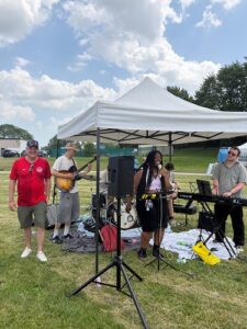 Small outdoor band performing under a white canopy in a grassy park, with guitarist, drummer, keyboardist, and a vocalist at the front.