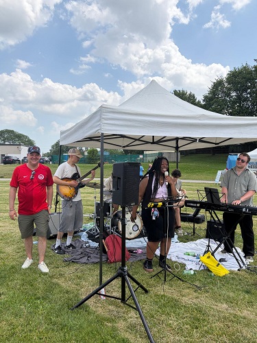Small outdoor band performing under a white canopy in a grassy park, with guitarist, drummer, keyboardist, and a vocalist at the front.