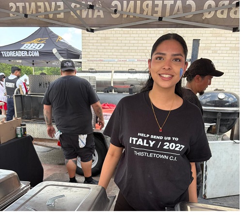 Smiling woman wearing a black 'Help send us to Italy 2027' T-shirt at an outdoor barbecue booth, with grills and volunteers visible in the background.