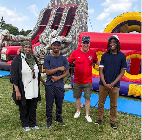 Four people standing together outdoors in front of a large inflatable slide and obstacle course at a sunny event, posing for the photo.